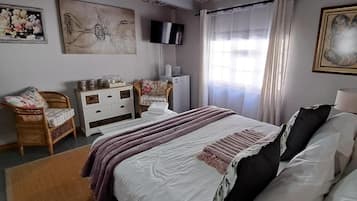 Une chambre à coucher comprenant un lit, une commode, une chaise, un téléviseur et une fenêtre avec des rideaux.