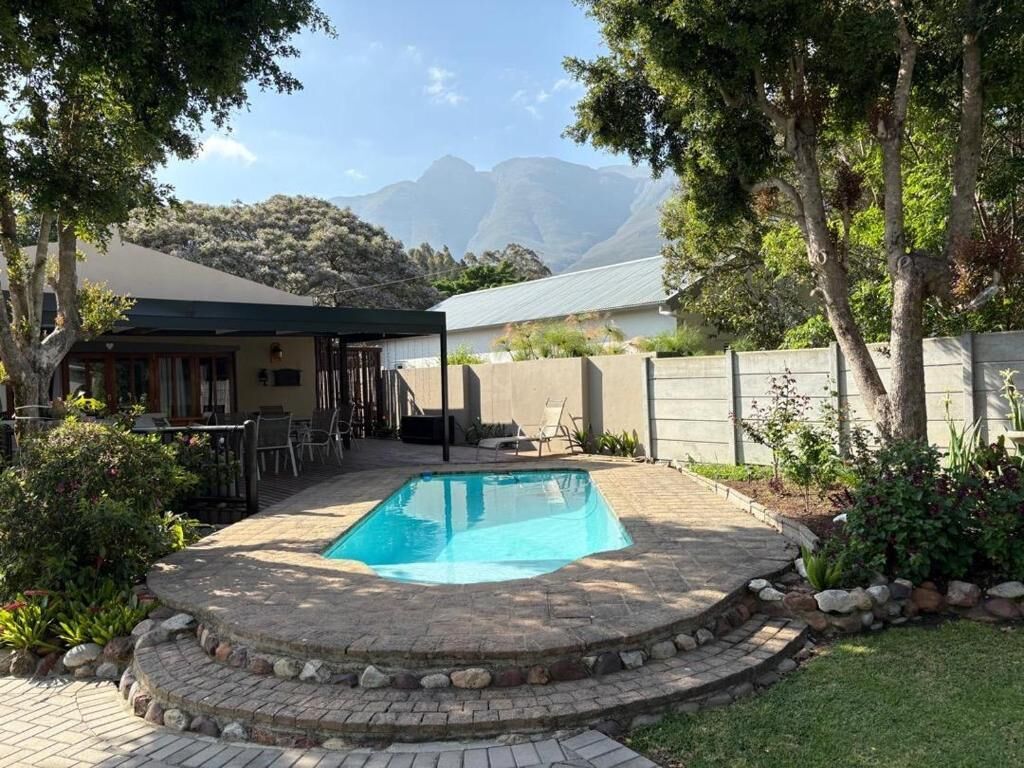 A swimming pool with a stone border, surrounded by a patio area with chairs and tables, and a view of mountains in the background.
