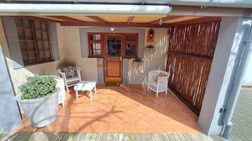 A covered patio area with a tiled floor, two white wicker chairs, a small table, and a potted plant.