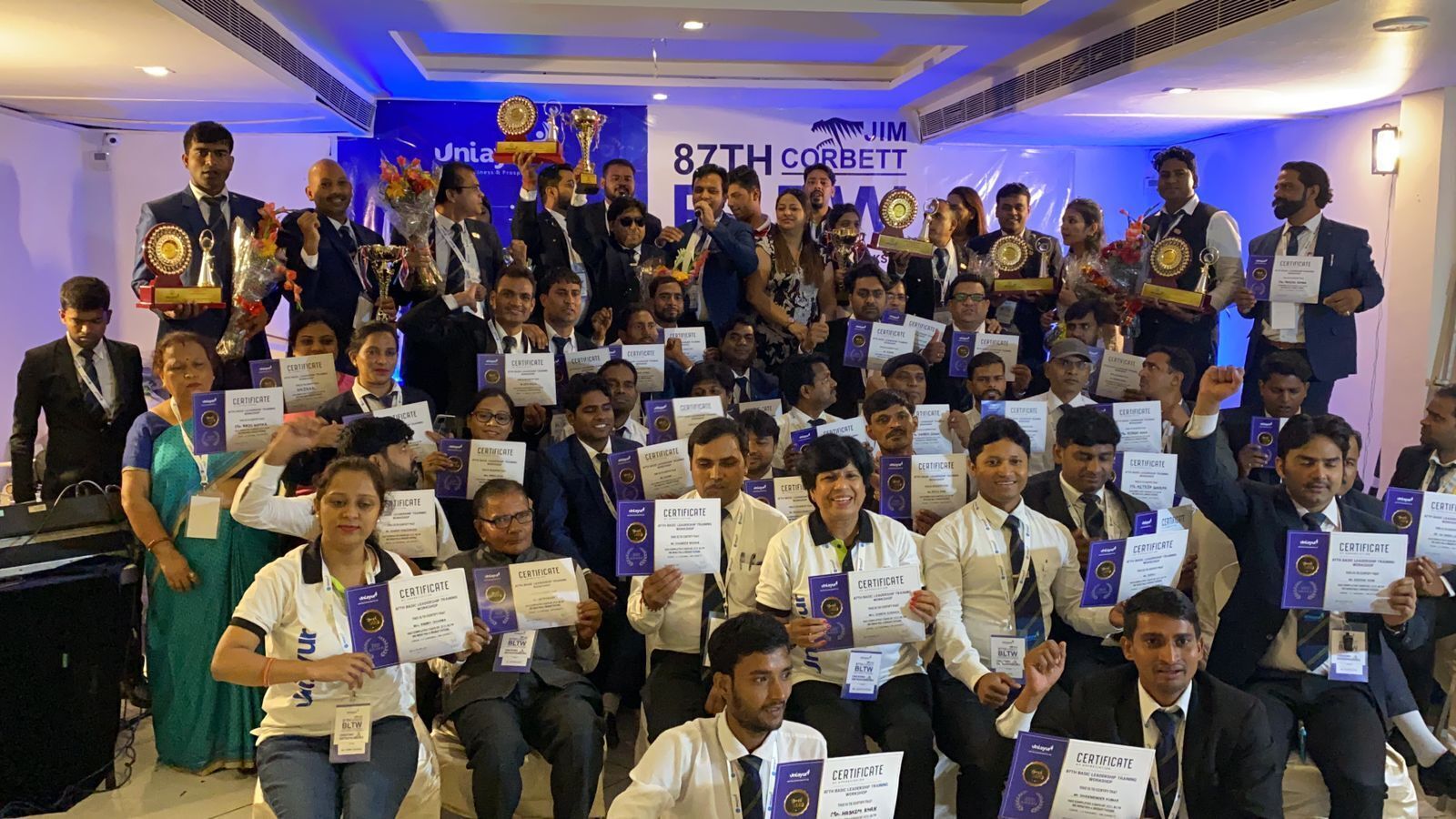 Un grupo de personas sosteniendo certificados y trofeos en un evento interior, con fondo iluminado en azul y un cartel con texto.