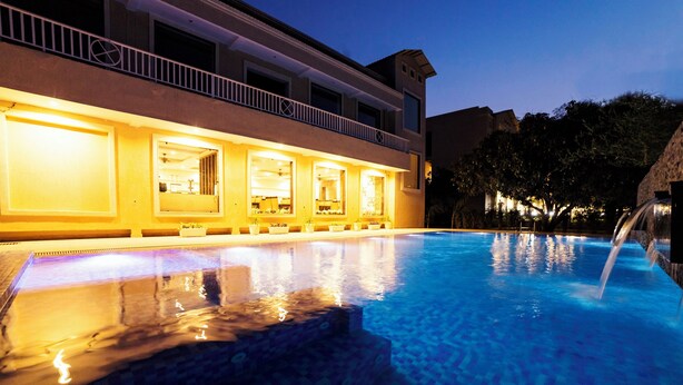 A swimming pool with a waterfall feature, a building with multiple windows, and a balcony with railings. - La Savanna Resort And Spa By DL Hotels And Resorts (Ramnagar)