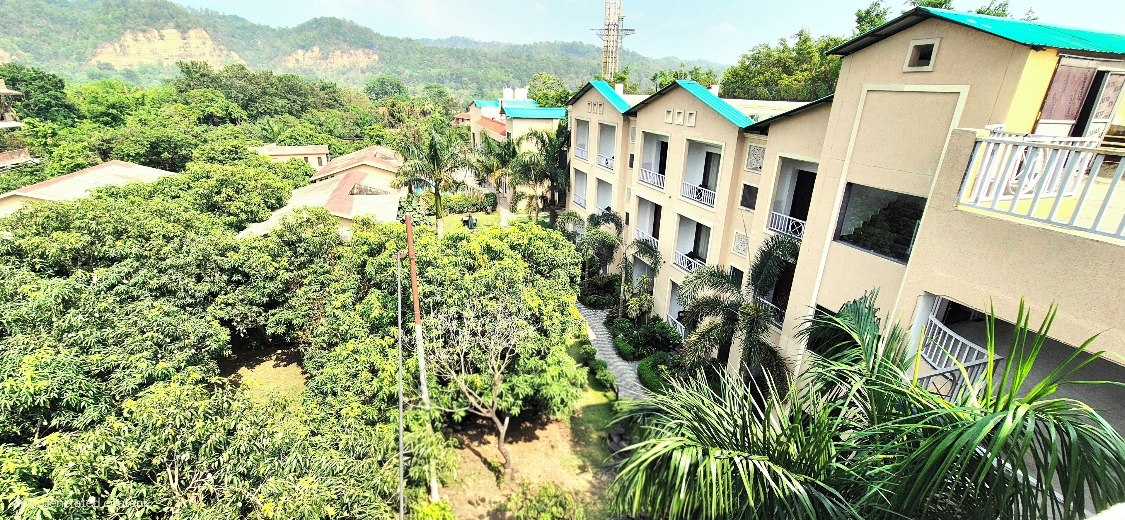 A multi-story building with balconies, surrounded by lush greenery and hills in the background.