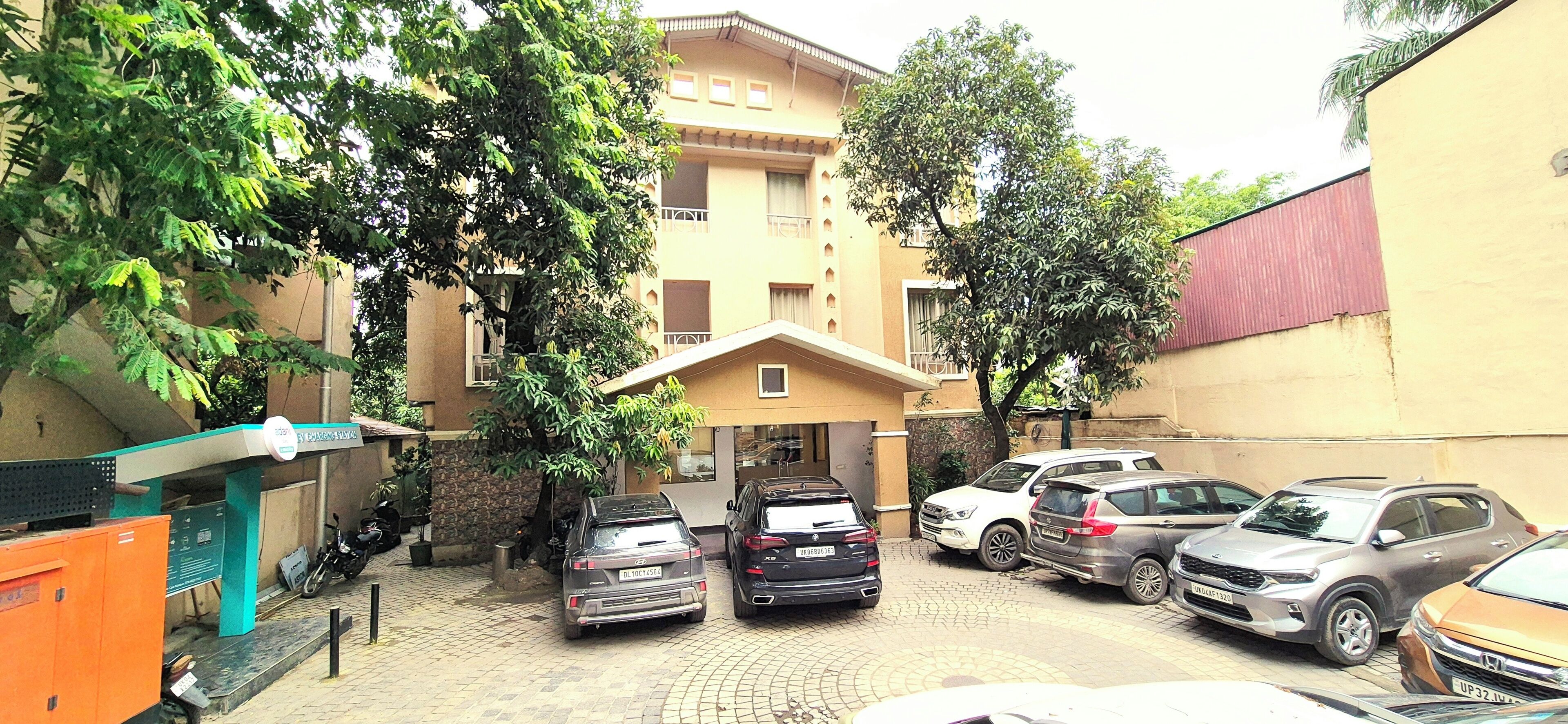 A parking area with multiple cars, a building with balconies, and a tree in the foreground.