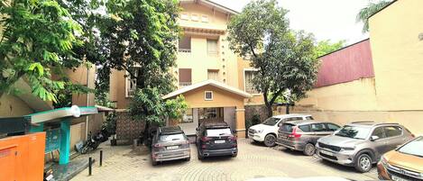 A parking area with multiple cars, a building with balconies, and a tree in the foreground.