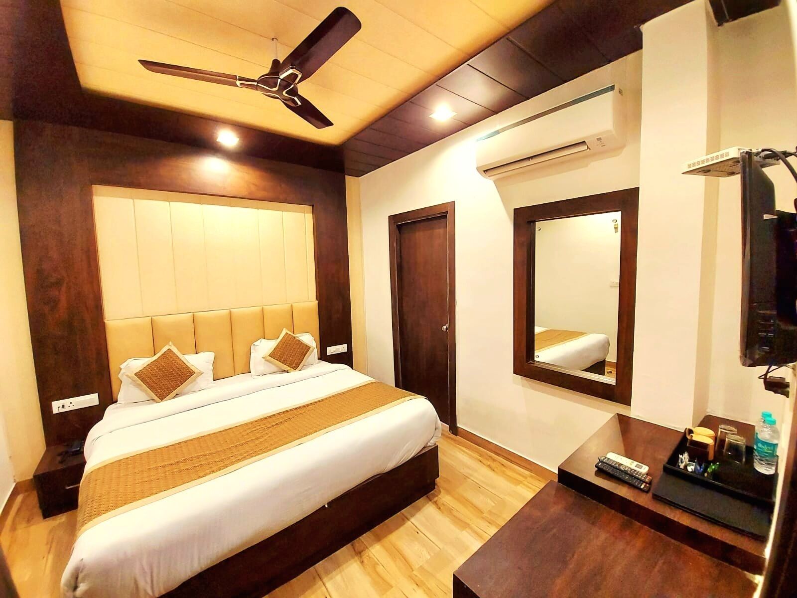 A hotel room with a bed, a ceiling fan, a mirror, a desk, and a TV.
