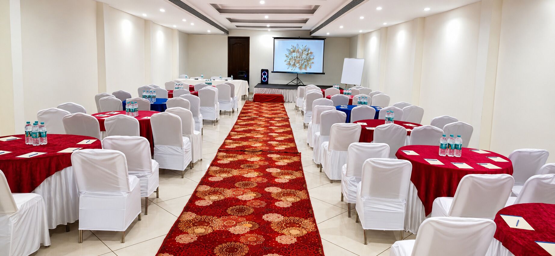 Un salón de conferencias con filas de mesas, sillas, un pasillo con alfombra y una pantalla mostrando 'WELCOME'.