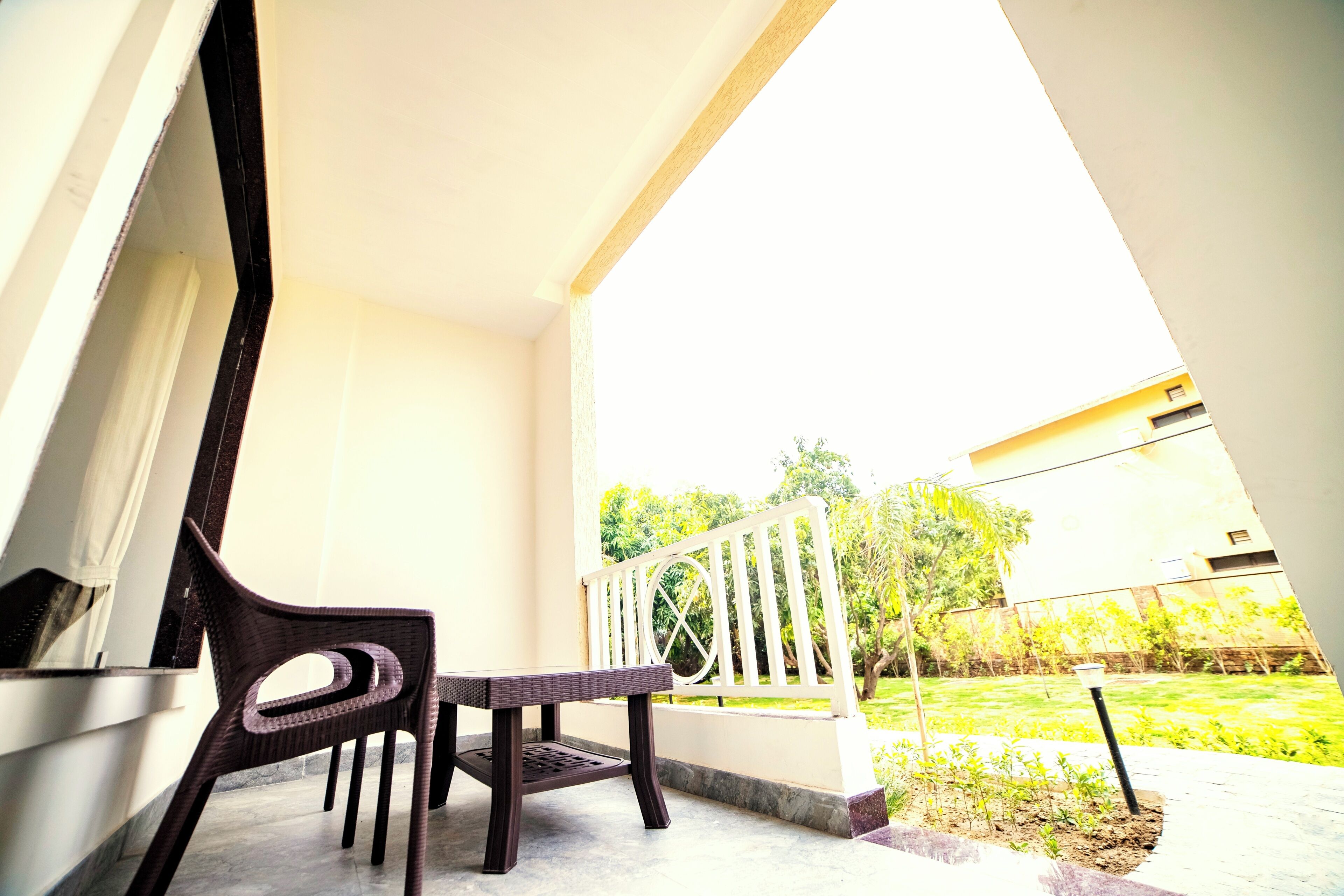A balcony with a chair and table, a white railing, and a view of a garden and building in the background.