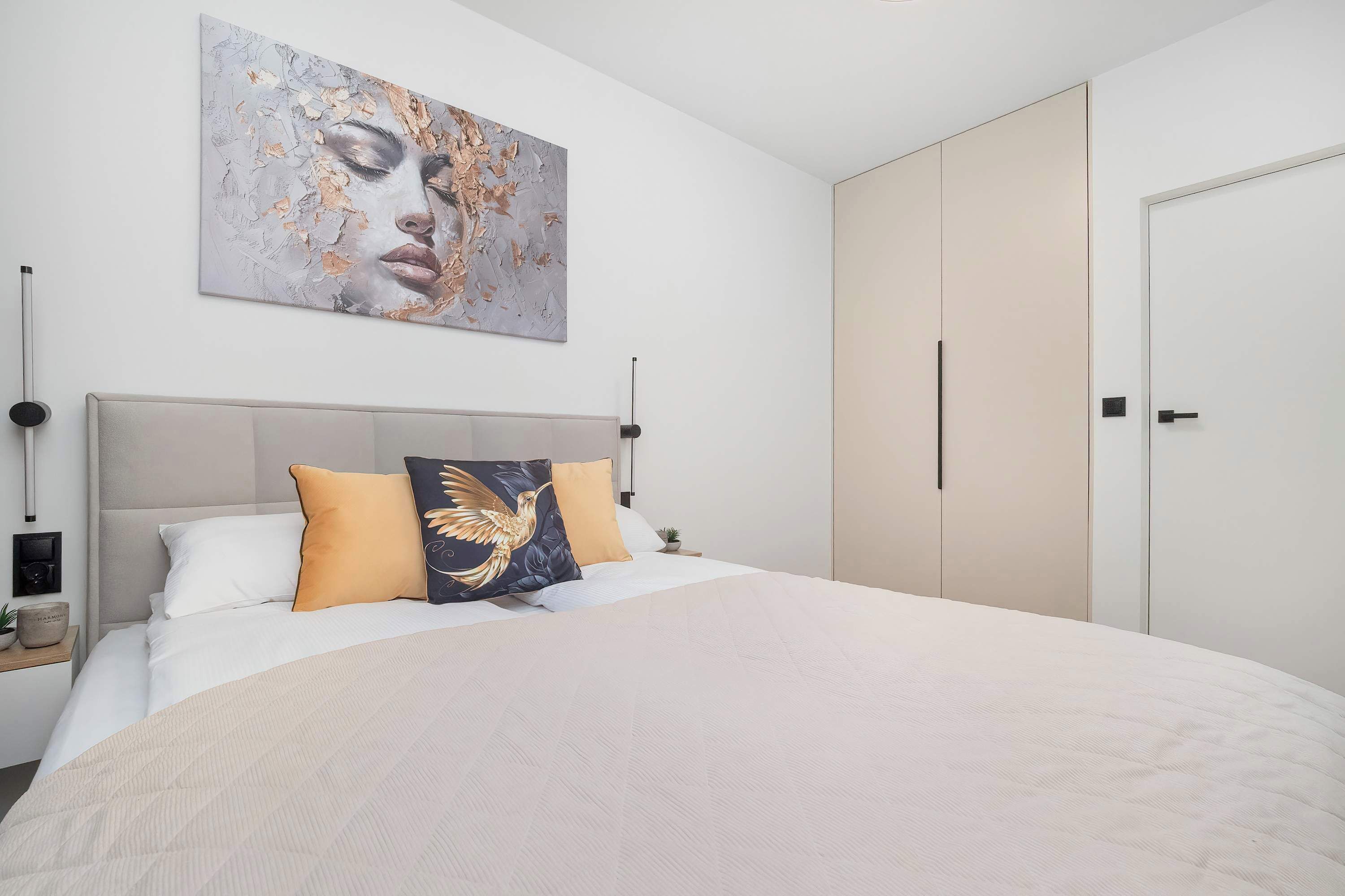 Een slaapkamer met een groot bed, hoofdeinde, een kunstwerk aan de muur, een kledingkast en een nachtkastje met een lamp.