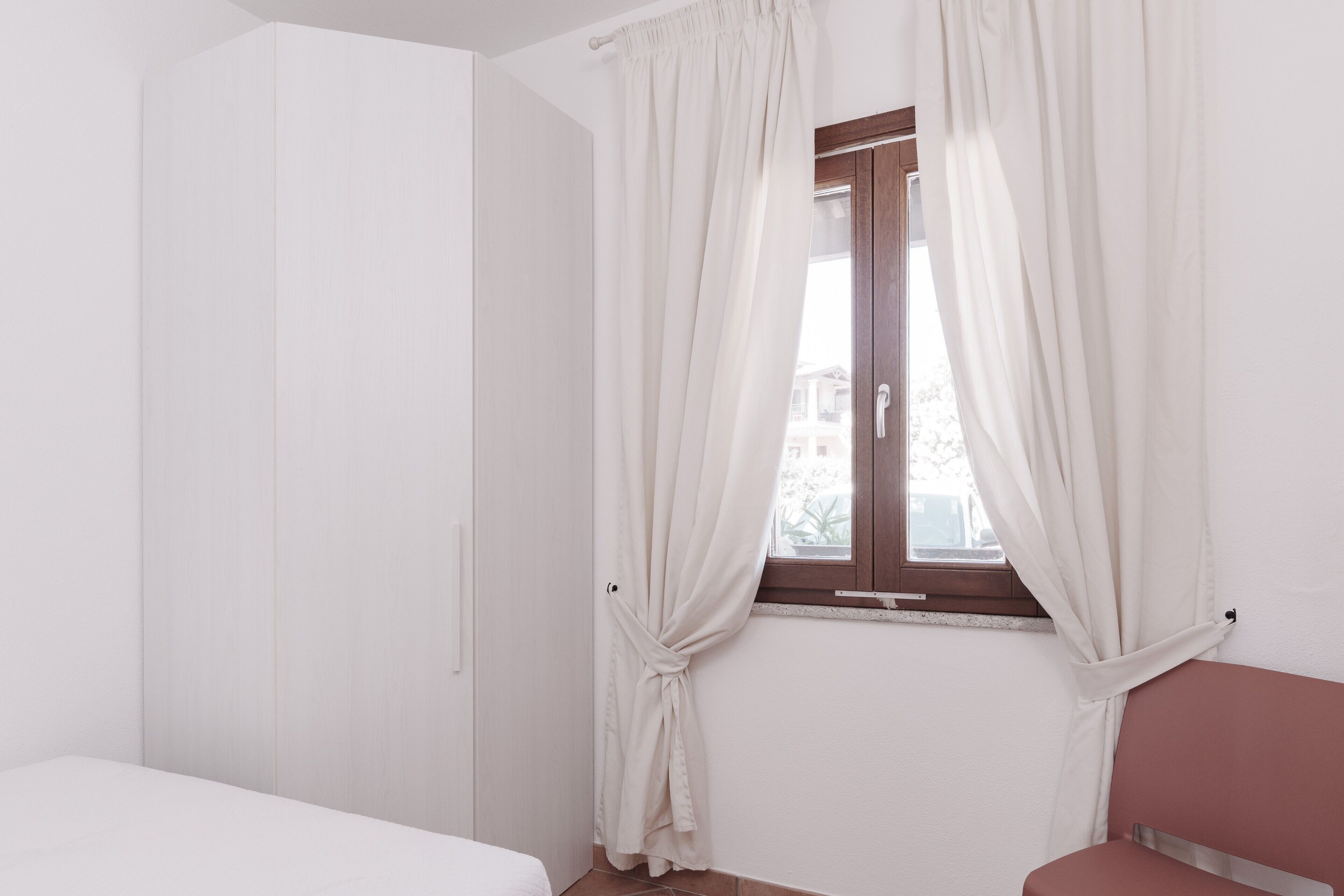 Una habitación con cama, armario, cortinas en la ventana y una silla.