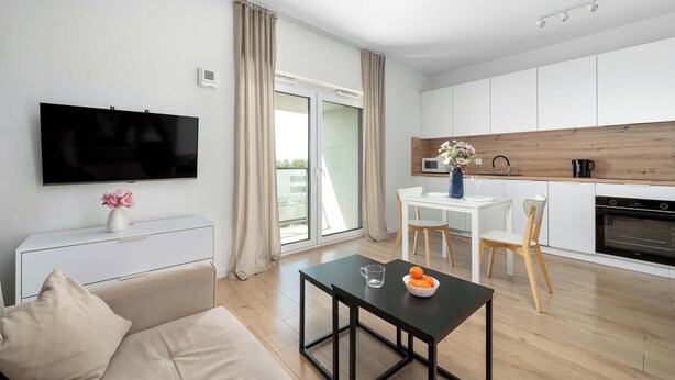 Una sala moderna con una televisión de pantalla plana, un sofá, una mesa de comedor con sillas, una zona de cocina con mostrador de madera y una puerta corrediza de vidrio que da a un balcón.