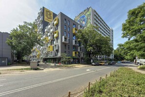 Ein mehrstöckiges Gebäude mit hervorgehobenem Logo an der Fassade, Balkonen und einer Straßenansicht mit parkenden Autos.
