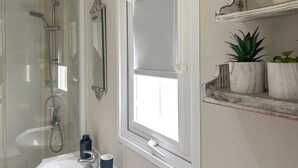 Une salle de bain avec une douche à parois de verre, un lavabo blanc, un miroir et une fenêtre. Des plantes en pot sont disposées sur une étagère.