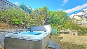 A hot tub in a backyard with a wooden deck, surrounded by greenery and a house in the background.