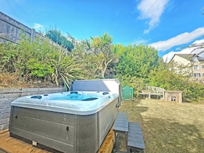 A hot tub in a backyard with a wooden deck, surrounded by greenery and a house in the background. - 2 bedroom accommodation in Paignton (Paignton)