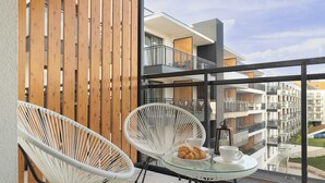 Un balcon avec deux fauteuils en osier blancs, une table en verre sur laquelle se trouvent une assiette de croissants et une tasse, et une vue sur des immeubles d’appartements modernes.