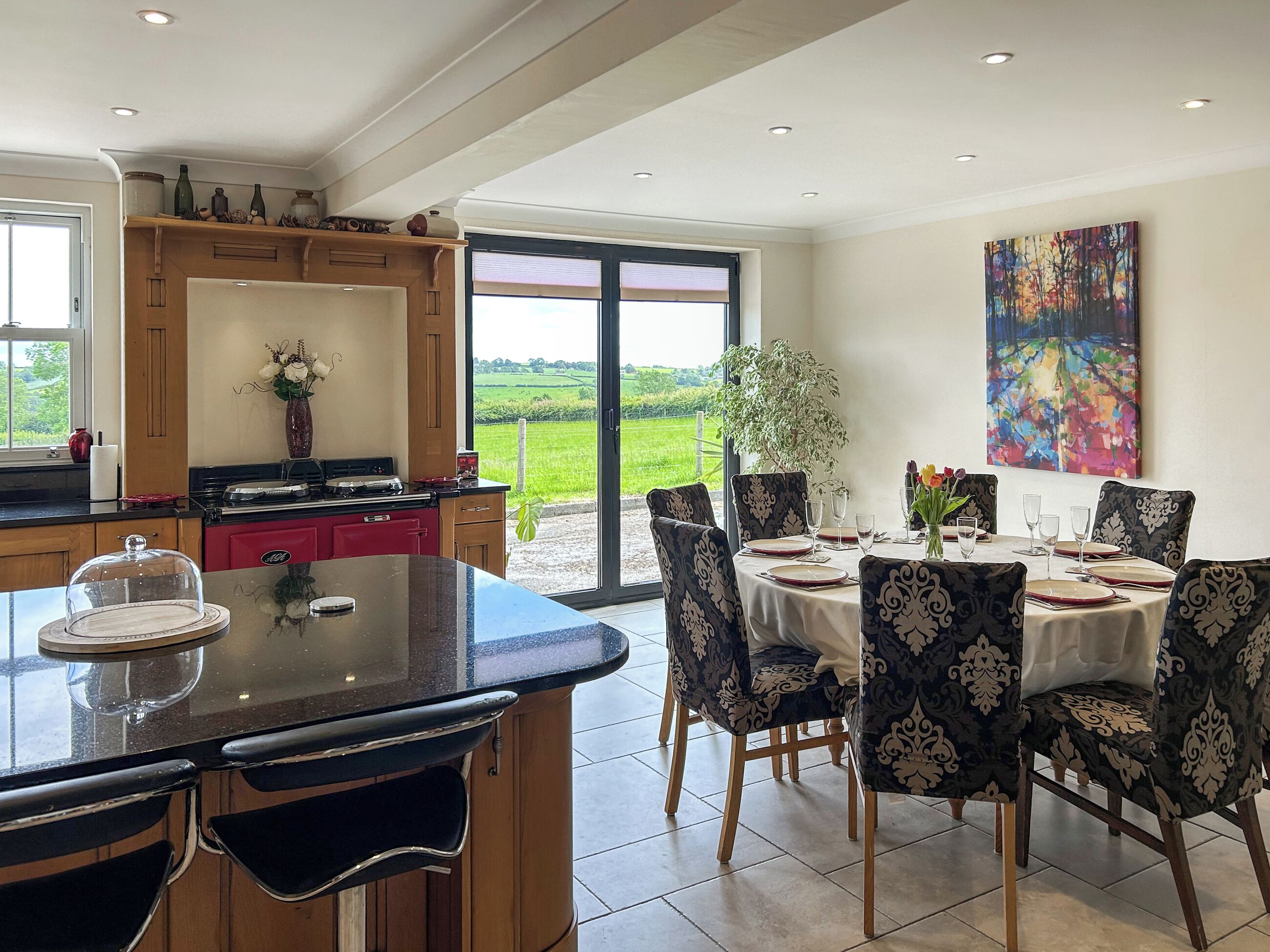 Un coin repas avec une table dressée pour quatre personnes, entouré de chaises à motifs, un espace cuisine avec une cuisinière et des placards, et une grande fenêtre offrant une vue sur un paysage verdoyant.