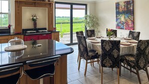Ein Essbereich mit Tisch für vier Personen, umgeben von stuhligen Sitzgelegenheiten mit Muster, ein Küchenbereich mit Herd und Schränken sowie ein großes Fenster mit Blick auf eine grüne Landschaft.