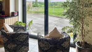 Ein Zimmer mit einer großen Schiebetür und Blick auf ein grünes Feld sowie einen Holzzaun. Zwei gemusterte Stühle und ein Blumentopf sind ebenfalls vorhanden.