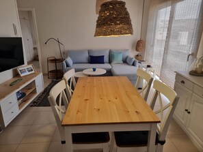 Un moderno salón con una mesa de comedor de madera, sillas blancas, un sofá con cojines azules, una lámpara colgante tejida y un televisor sobre un mueble.