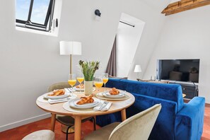 A dining area with a round table, chairs, plates of food, glasses of orange juice, and a television in the background. - Charming 1BR Hidden Gem in Le Marais (Paris)