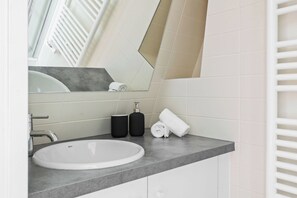 A bathroom with a white sink, a mirror, a towel, and two black bottles. - Charming 1BR Hidden Gem in Le Marais (Paris)