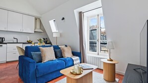 En moderne stue med en blå sofa, et træbord, en lampe og udsigt over byen gennem et vindue.