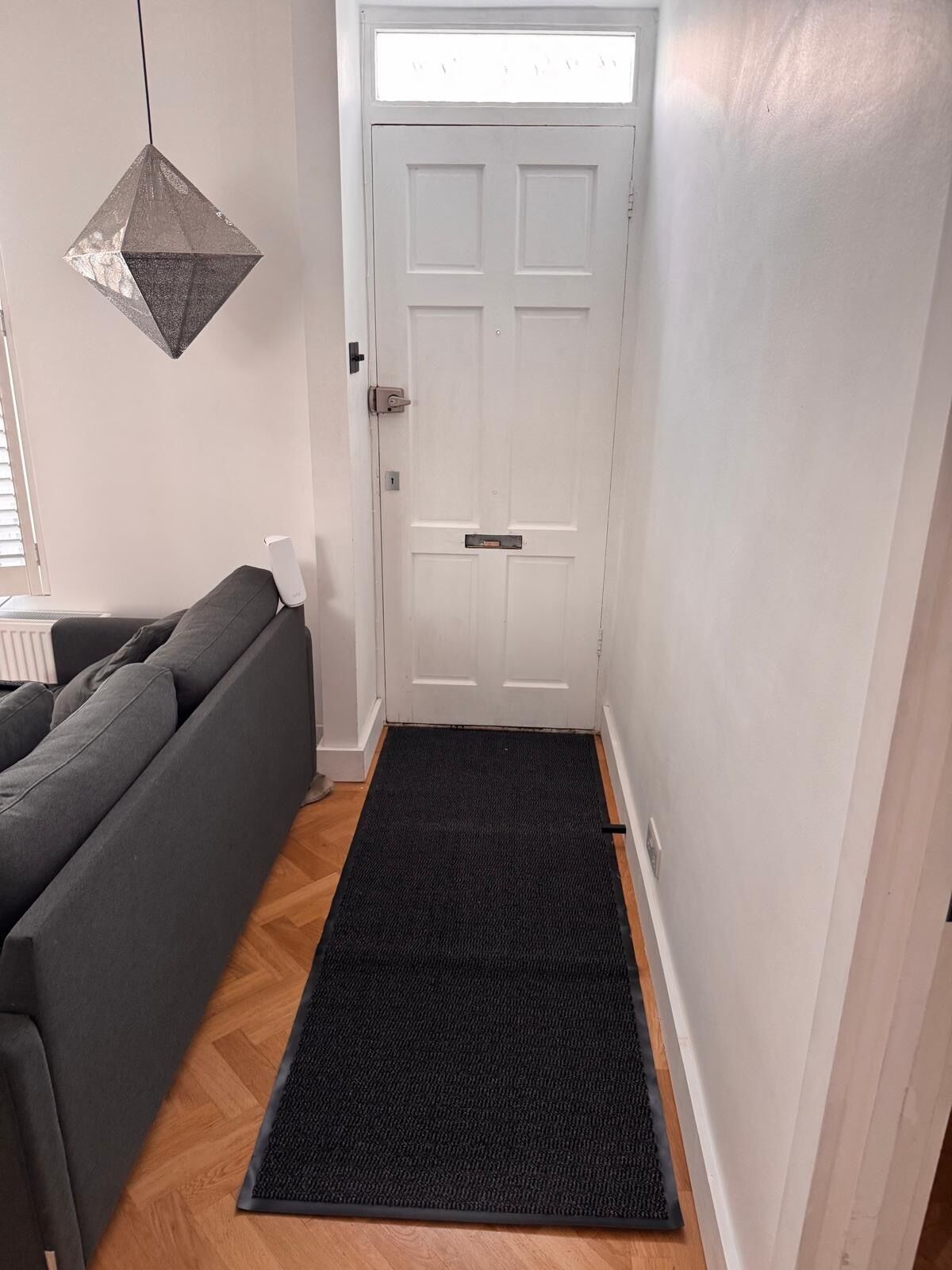 En hall med en vit dörr, en svart dörrmatta, en grå soffa och en hängande lampa.