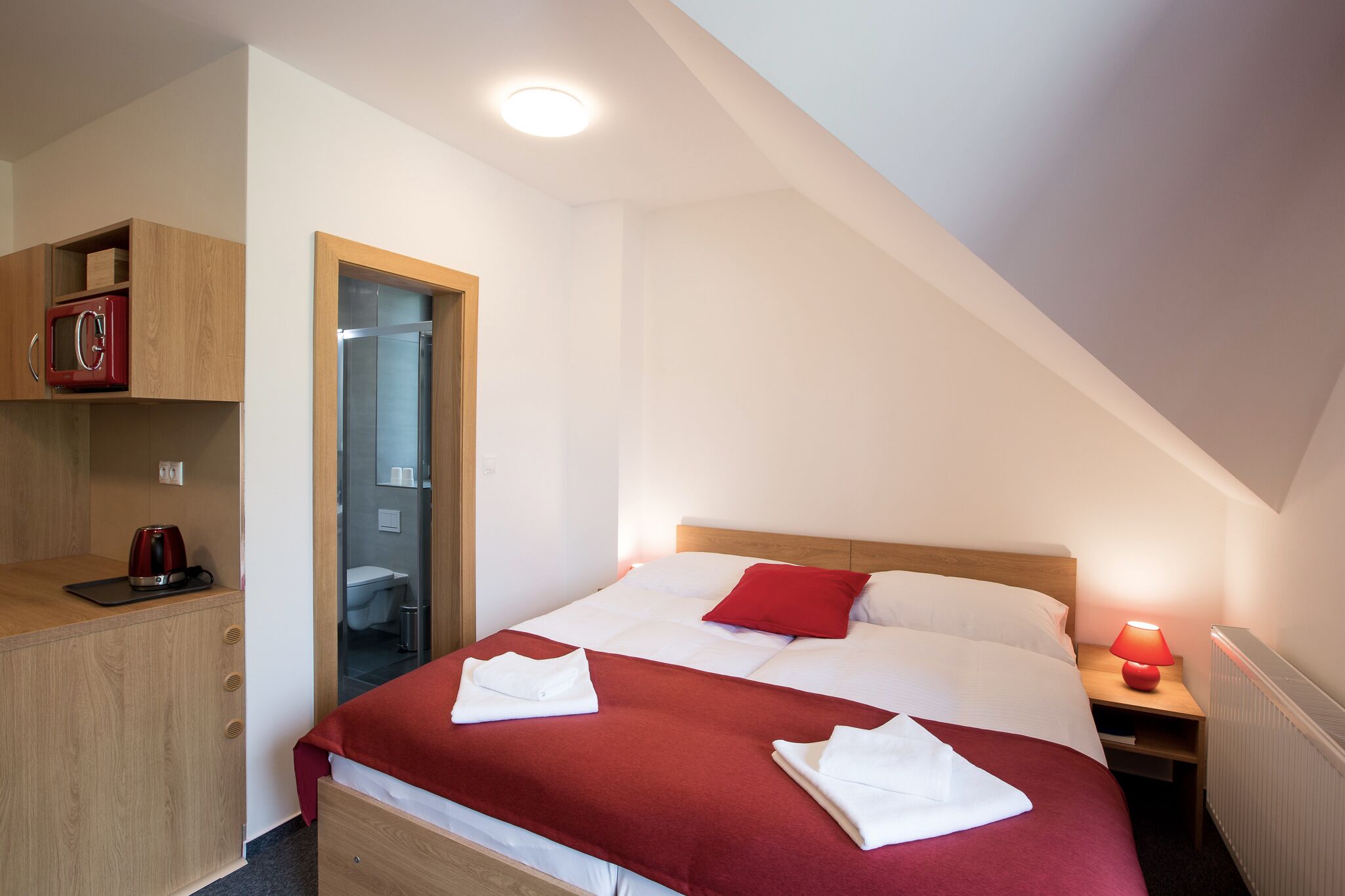 A bedroom with a bed, a nightstand, a red lamp, a wooden wardrobe, and a bathroom visible through a glass door.