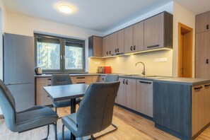 A modern kitchen with wooden cabinets, a dining table with chairs, and a window with blinds. - Vila DEDO (Vysoké Tatry)