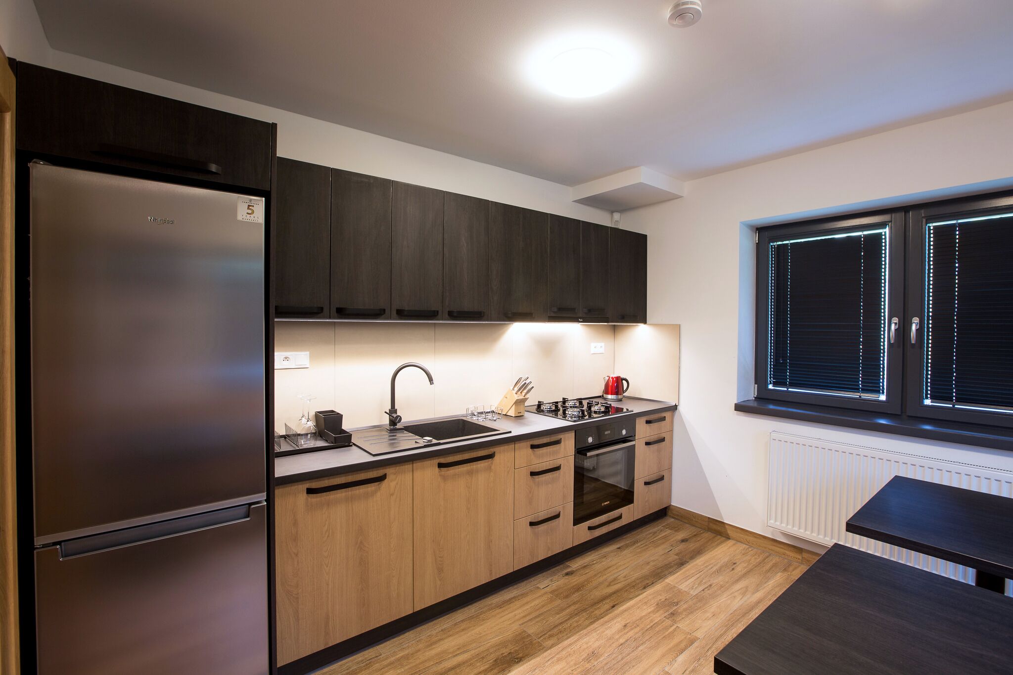 Una cocina moderna con un refrigerador de acero inoxidable, armarios de madera, un lavamanos, una estufa y una ventana con persianas.
