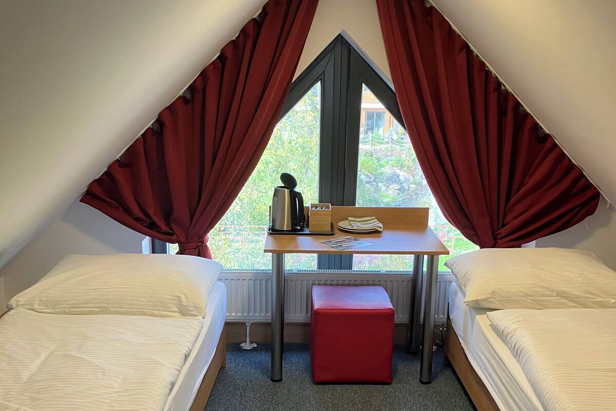 Una habitación pequeña y triangular con dos camas, un escritorio y una ventana con cortinas.