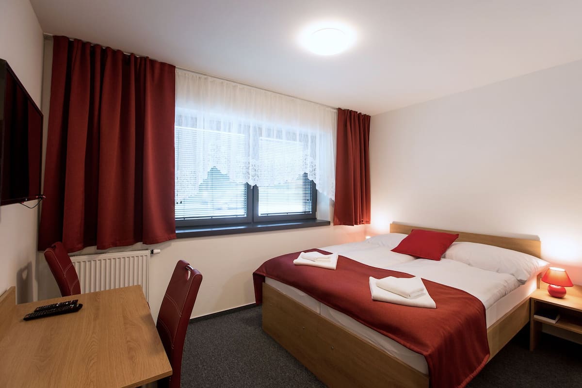 Una habitación de hotel con cama, escritorio, silla, ventana con cortinas y mesita de noche con lámpara.