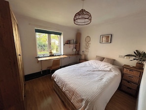 Ein Schlafzimmer mit einem großen Bett, einem Schreibtisch mit Stuhl, einem Fenster, einem Wandregal und einer Pendelleuchte.