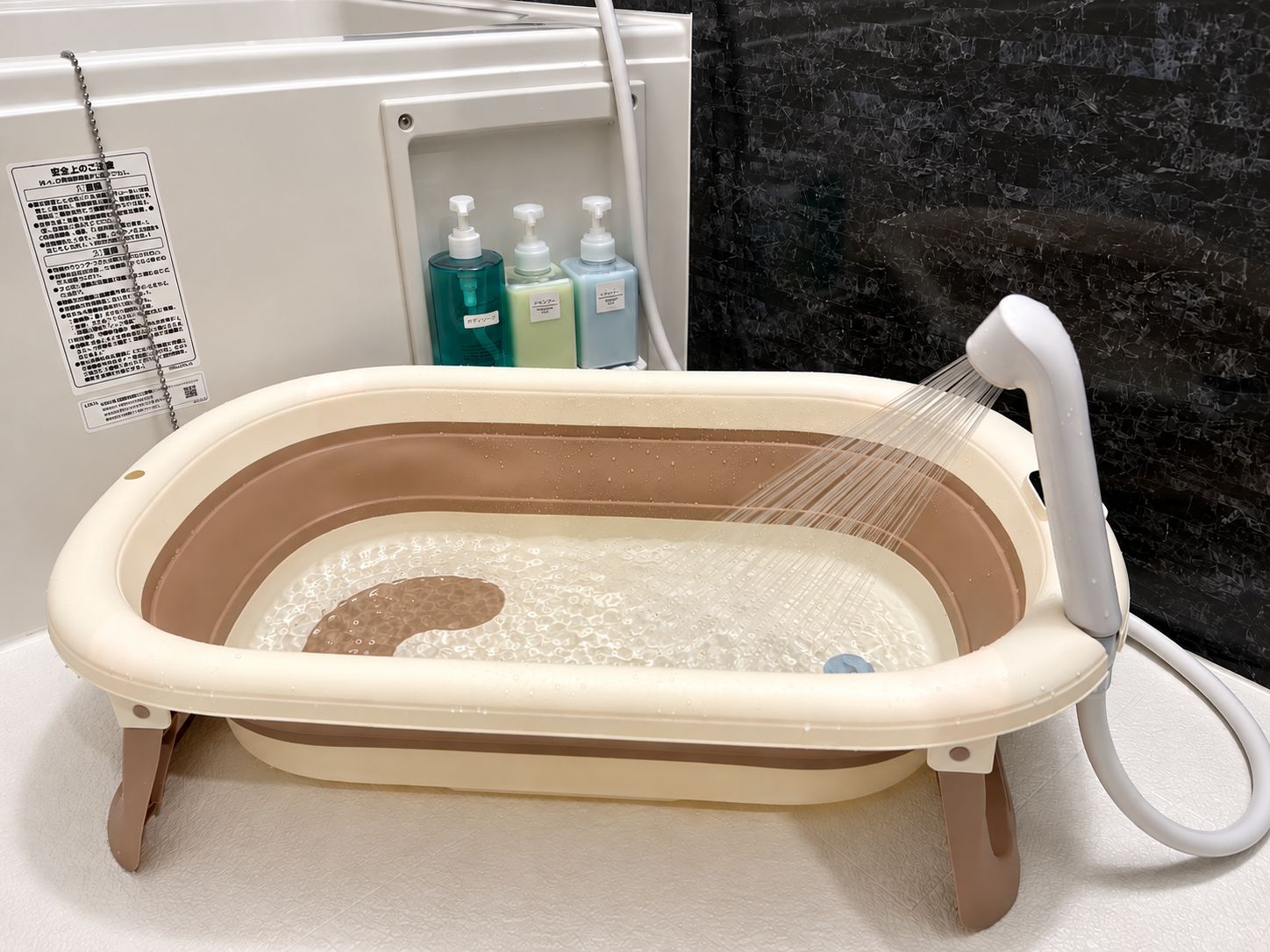 Una bañera portátil con cabezal de ducha, sobre una superficie blanca frente a una pared negra con textura.