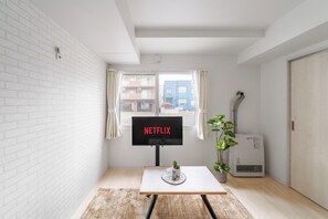 A room with a white brick wall, a window with curtains, a flat-screen TV displaying 'NETFLIX,' a small table with a plant, and a heater. - PREGARE 103 (Sapporoshi)