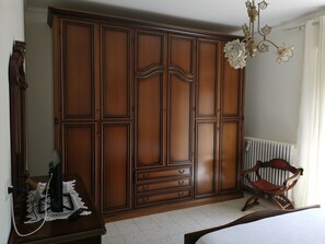 A bedroom with a large wooden wardrobe, a chair, a radiator, and a chandelier.