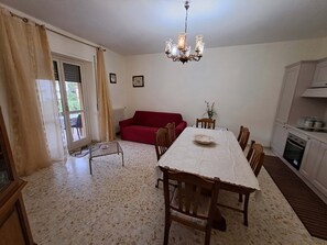 A dining area with a table, chairs, a red sofa, a chandelier, and a kitchen in the background.