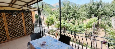 An outdoor balcony with a table and chairs, overlooking a garden and distant mountains.