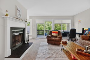 A living room with a fireplace, a sofa, a coffee table, a desk with a computer, and a view through large windows. - Wintergreen Retreat | Golf | Sleeps 12 | Pets OK (Wintergreen Resort)