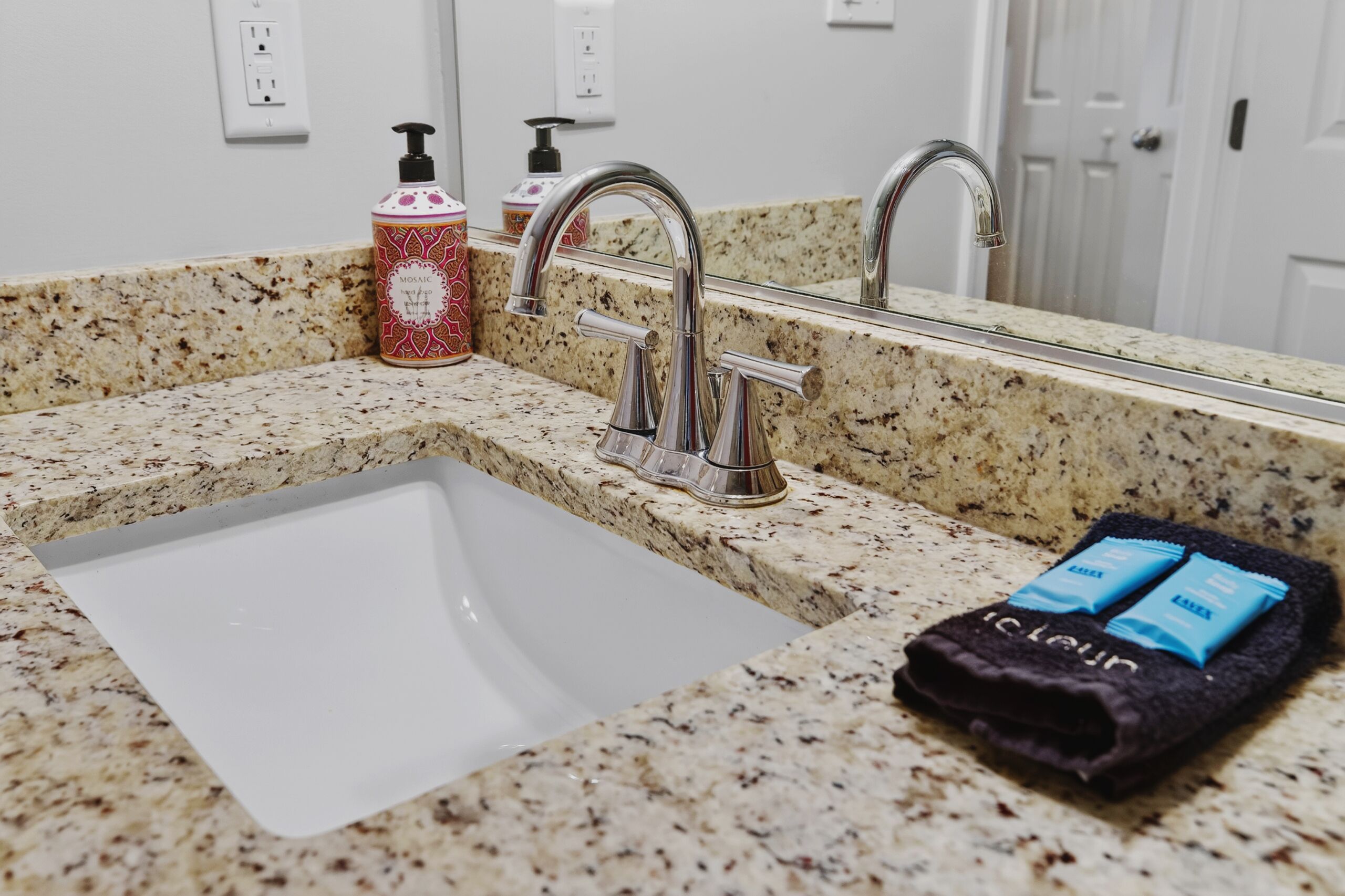 A bathroom with a granite countertop, a white sink, a mirror, a towel rack, and toiletries.