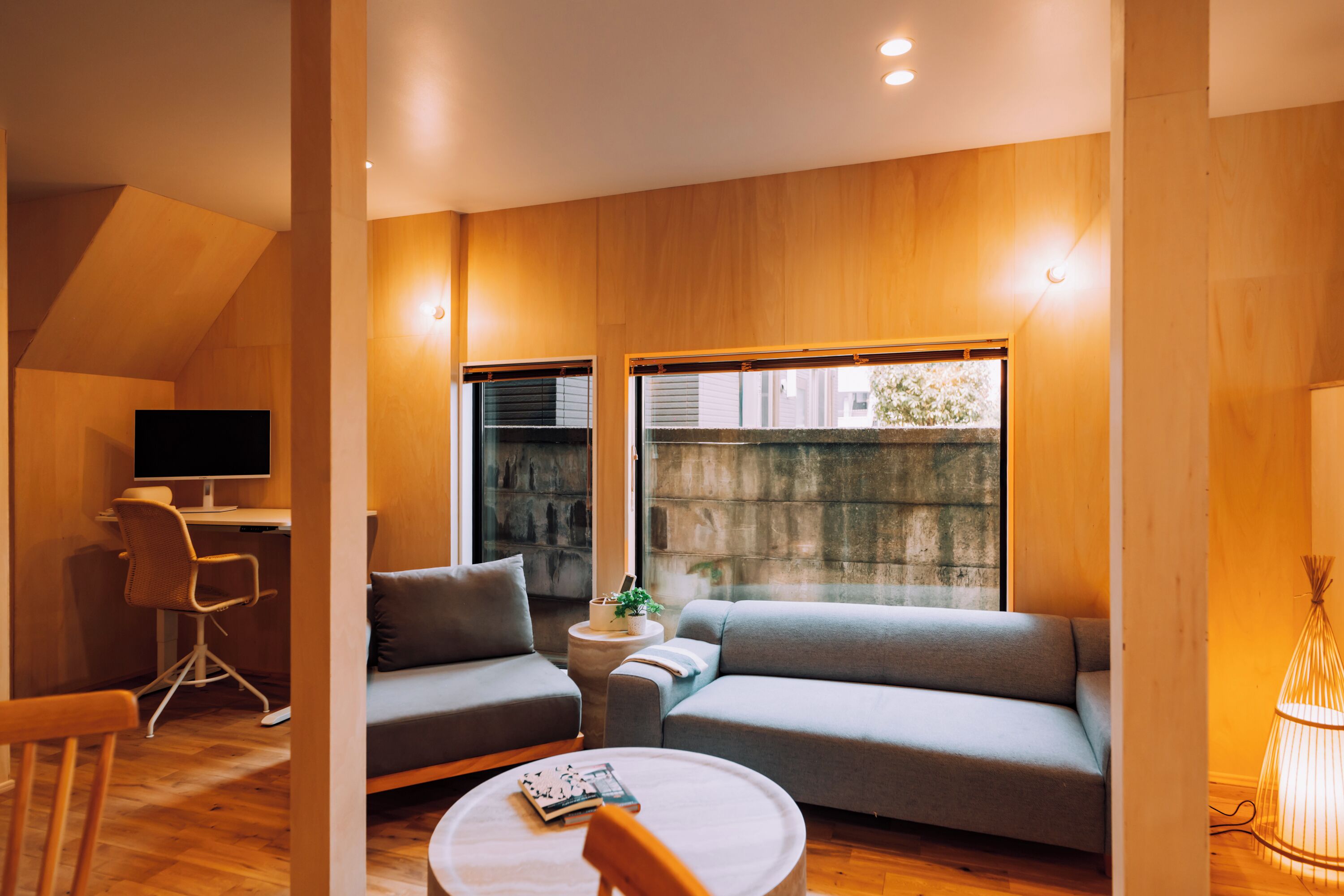 A modern living room with a sofa, a small round table, a chair, a desk with a computer, and a window with a view.