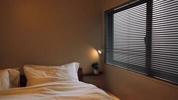 A bedroom with a bed, pillows, a bedside table with a plant, and a window with blinds.