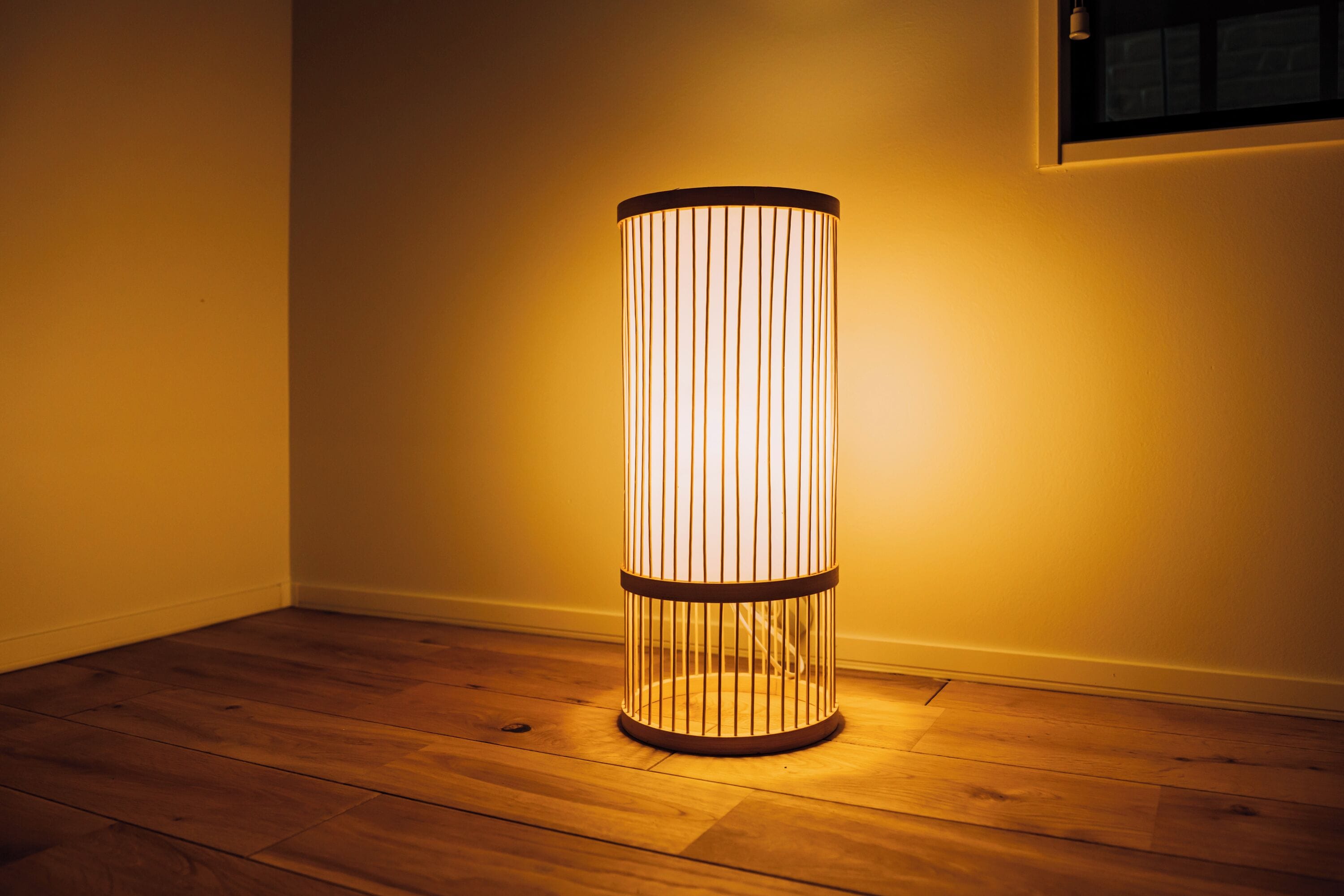 Een cilindrische vloerlamp met verticale strepen, staand op een houten vloer in een kamer met effen wanden.