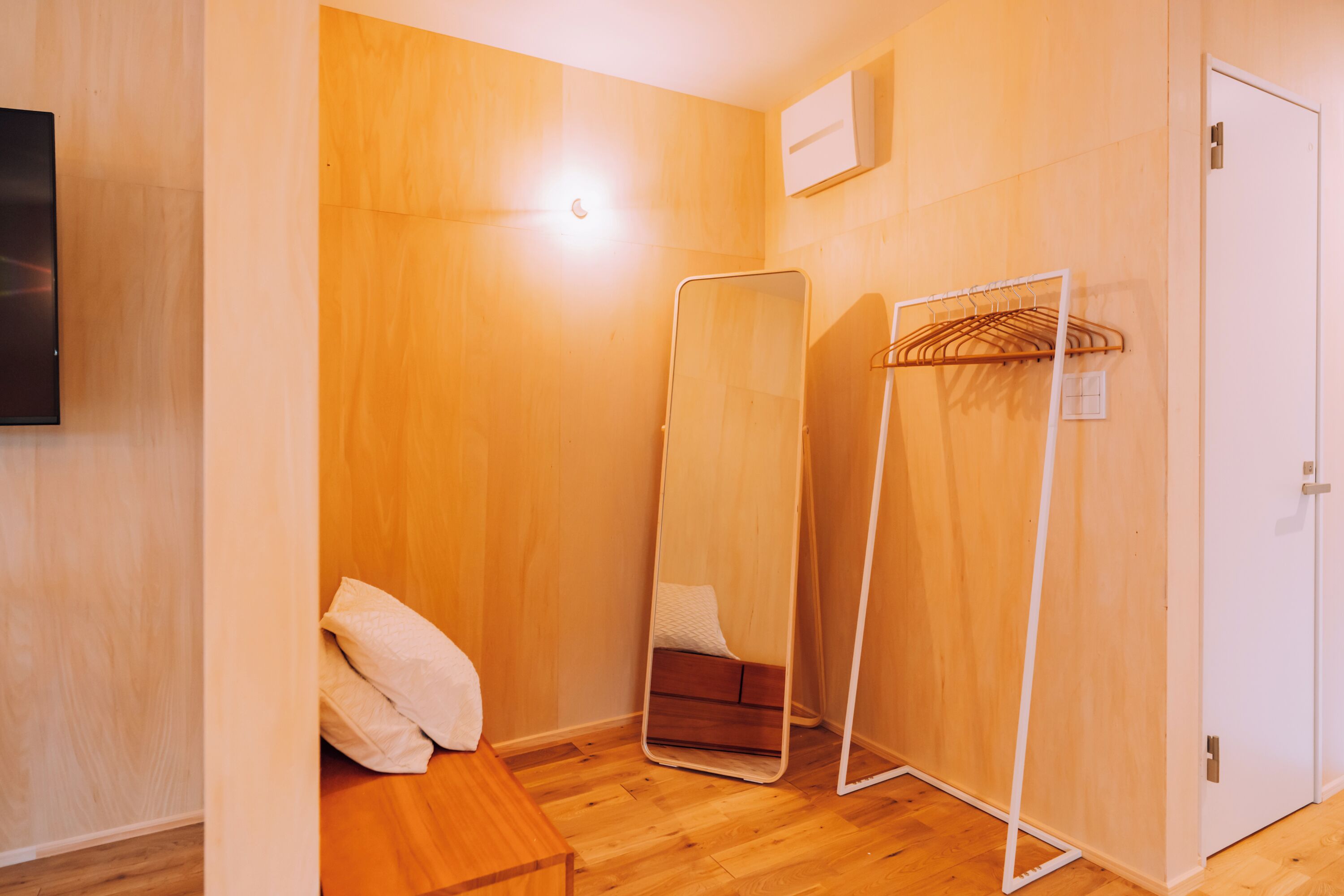 A room with wooden walls, a wooden bench, a mirror, a white door, and a wall-mounted air conditioning unit.