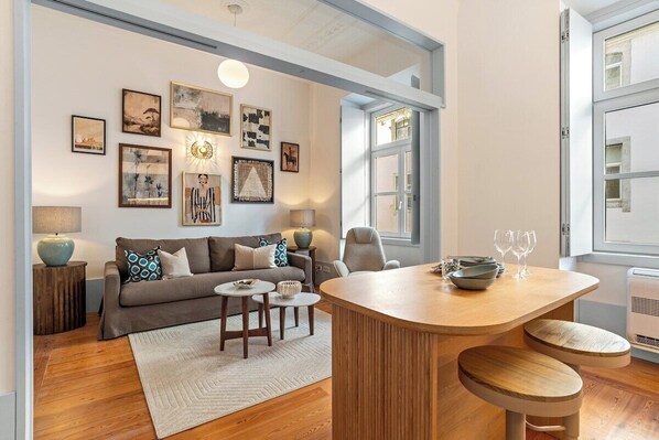 Ein modernes Wohnzimmer mit Sofa, Couchtisch und Sesseln, ein Essbereich mit Holztisch und Hockern sowie ein großes Fenster mit Aussicht.