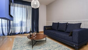 Ein modernes Wohnzimmer mit einem dunkelblauen Sofa, einem runden Couchtisch, einer Deckenleuchte und einem großen Fenster mit Vorhängen.