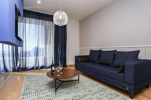 A modern living room with a dark blue sofa, a round coffee table, a chandelier, and a large window with curtains. - Status M Apartments WOW (Budva)