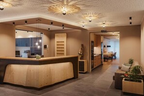 A modern interior with a wooden reception desk, a dining area, and a living space with a sofa and plants. - Hotel La Müda (Badia)