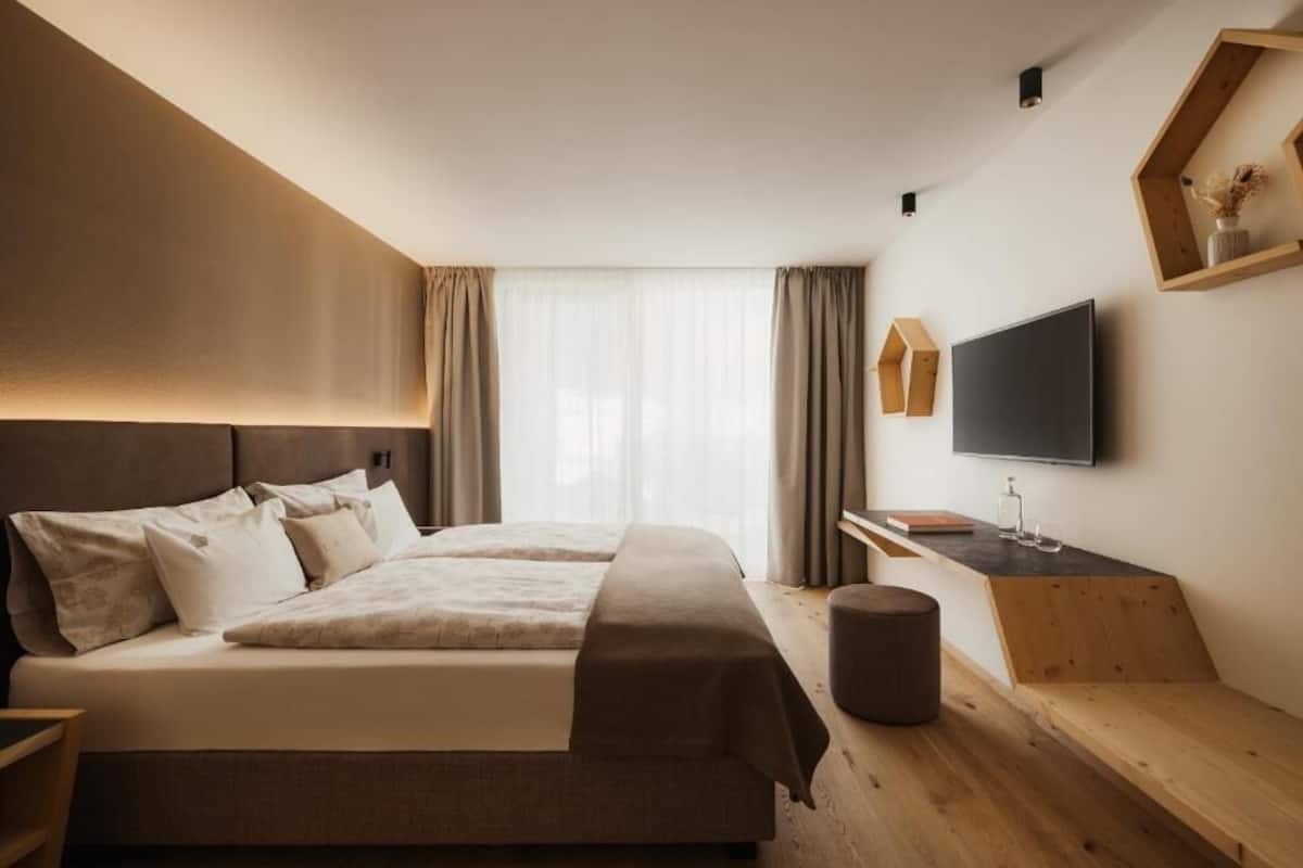 Una habitación de hotel moderna con una cama grande, un escritorio, un televisor de pantalla plana y un ventanal con cortinas.