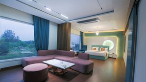 Een moderne hotelkamer met een hoekbank, een rond poefje, een kleine tafel, een bed met hoofdeinde, en een groot raam met uitzicht op de omgeving.